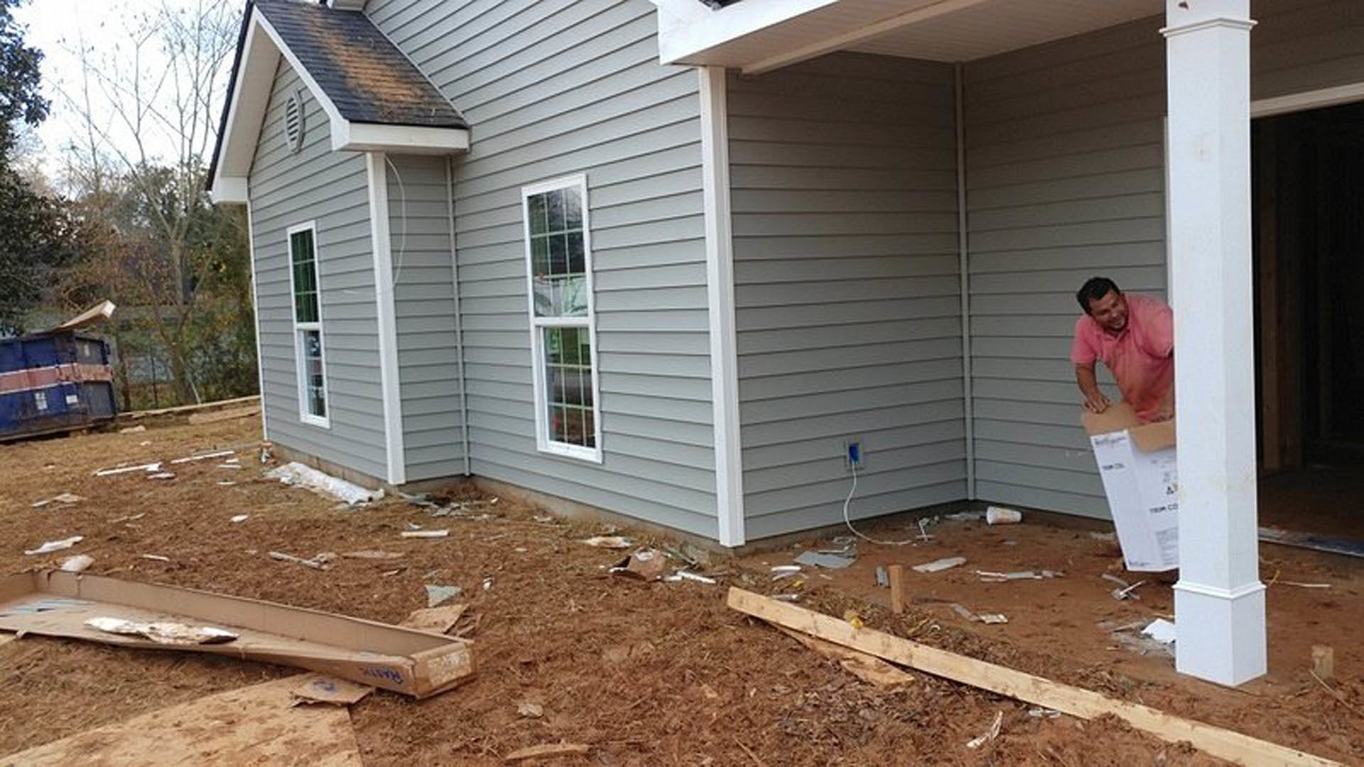 Partially built house with white-framed window, exposed siding, construction debris and metal scraps scattered on bare ground, man in pink shirt working nearby