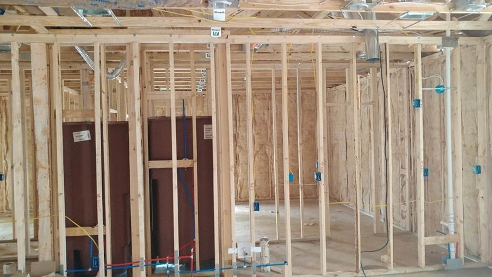 Exposed wood framing with beams and planks, construction materials including a white container with black text, blue rope, and labeled boxes inside unfinished residential structure