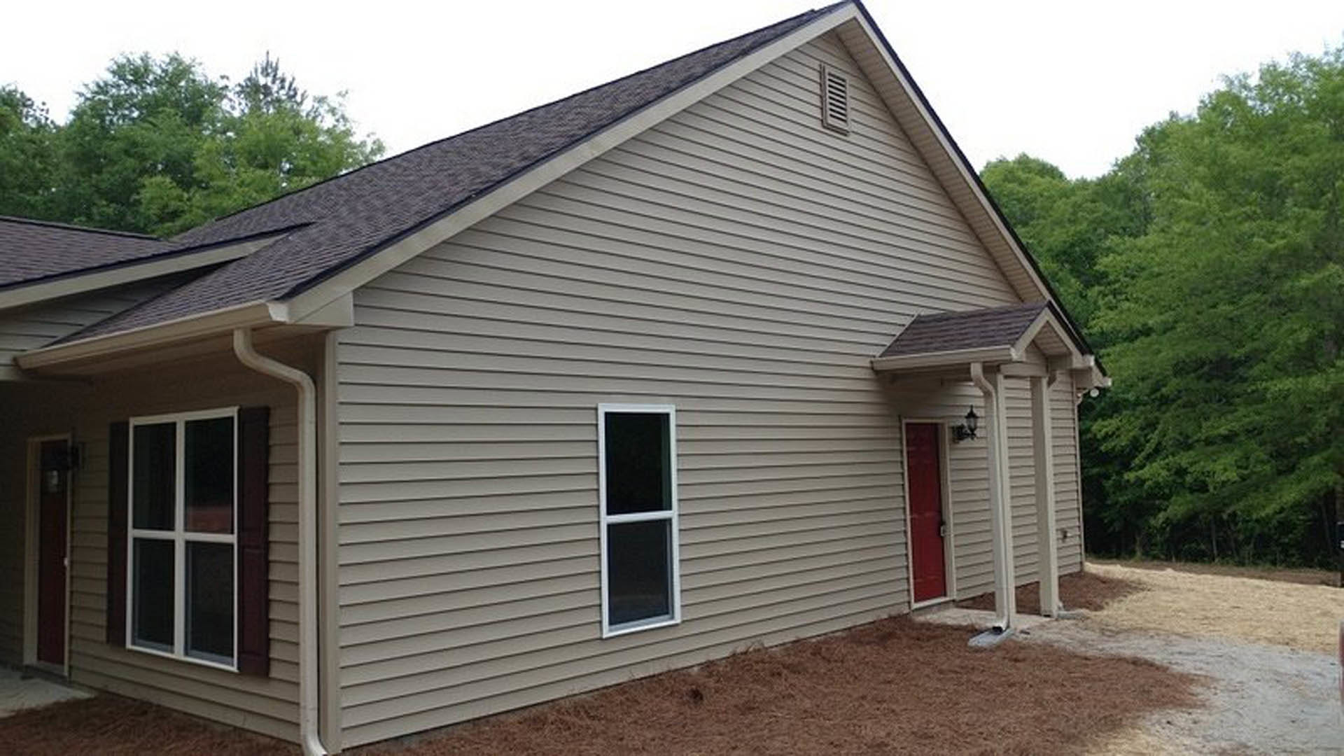 Two-story cottage-style home with gray siding, white-trimmed windows, prominent red front door, gabled roof, mature trees in background