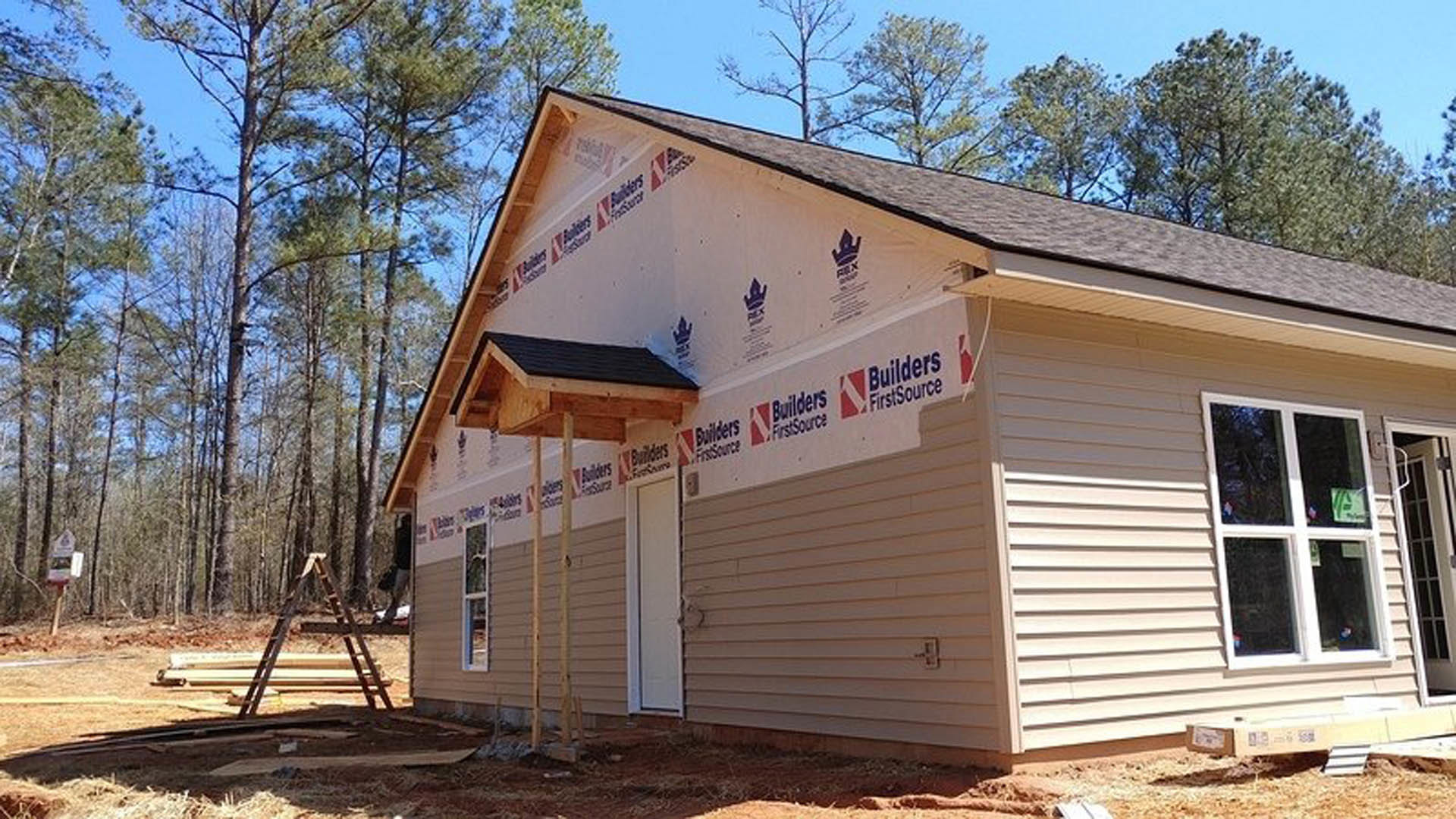Partially built home with exposed wooden framing, exterior siding, ladder leaning against wall, windows with construction signs, surrounded by trees and sky