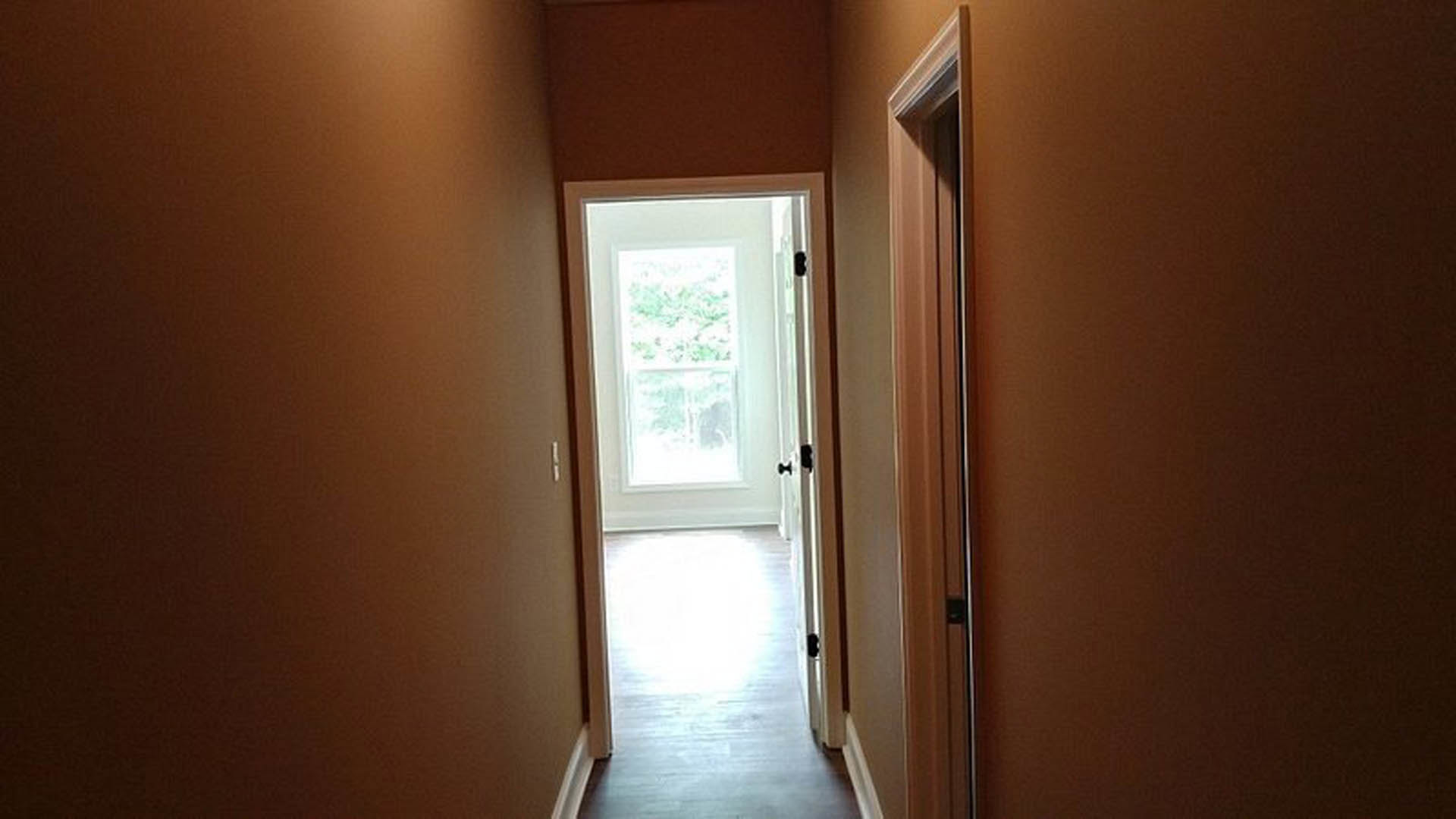 Open white door with brushed metal handle leading from hallway into sunlit room, grey plaster walls, light wood flooring, and ceiling with recessed lighting visible