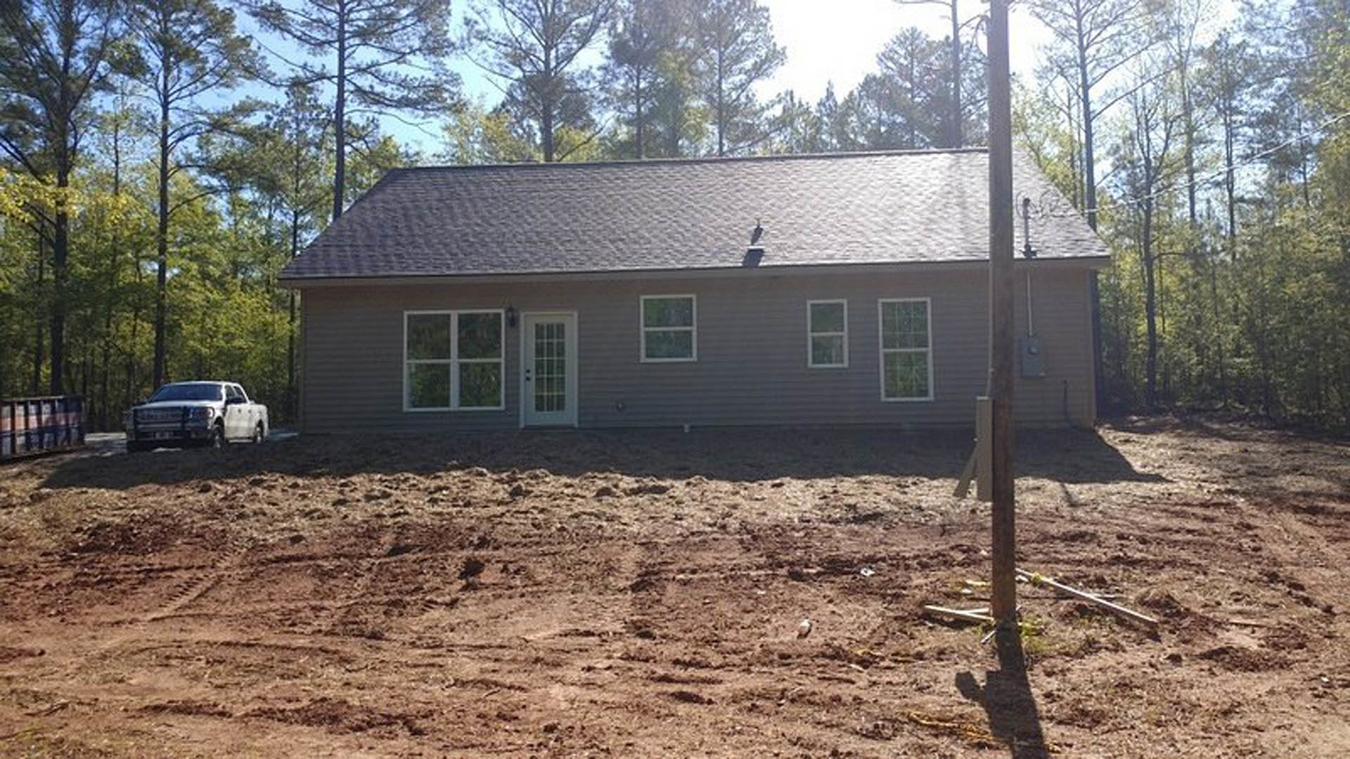 Two-story house with beige siding, white-framed windows, glass panel front door, fenced backyard, dirt field in foreground, white pickup truck parked on road, mature trees in