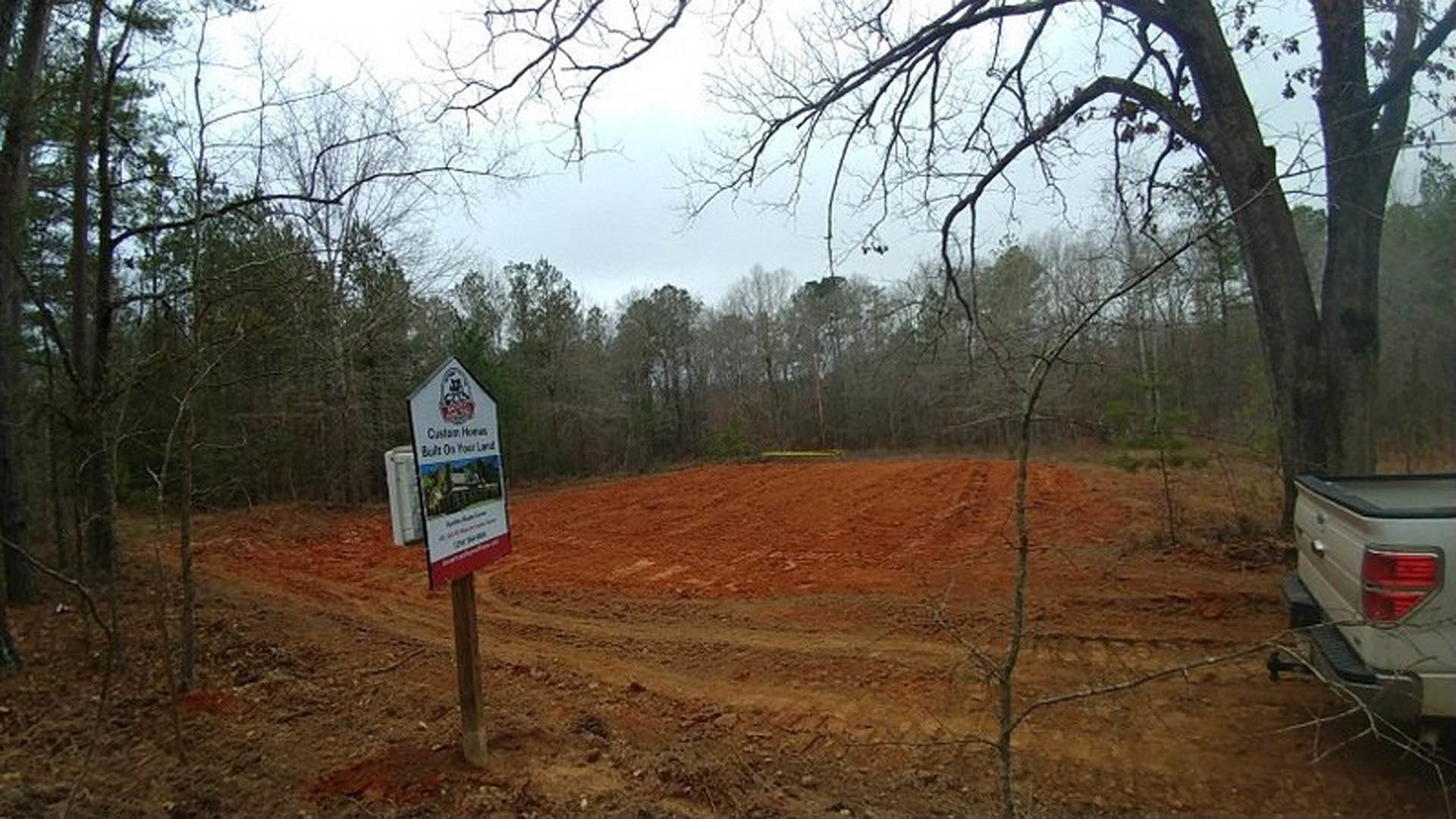 Wooden post sign in a dirt field with scattered grass, trees in the background, and partial view of a truck tail light