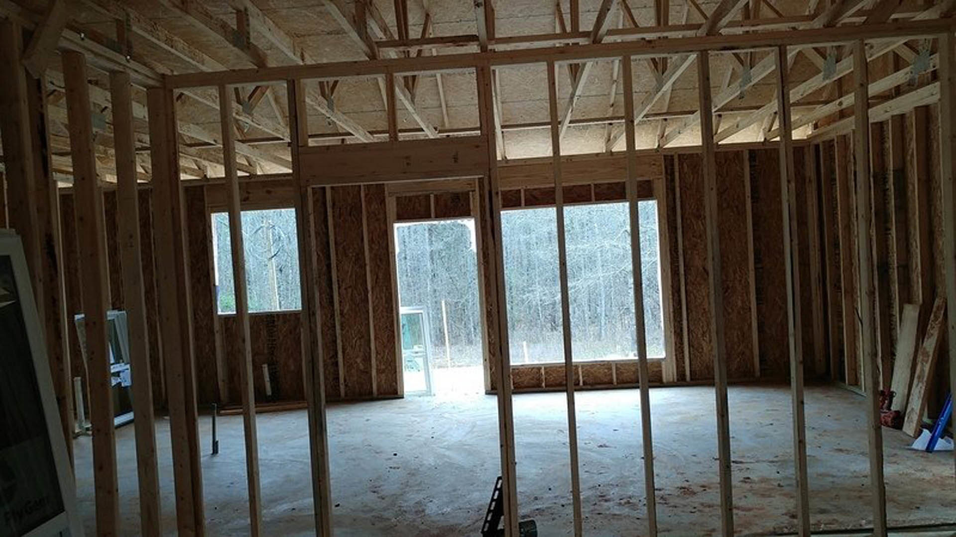 Sunlit room with large windows, wooden door, exposed ceiling beams, and light wood flooring