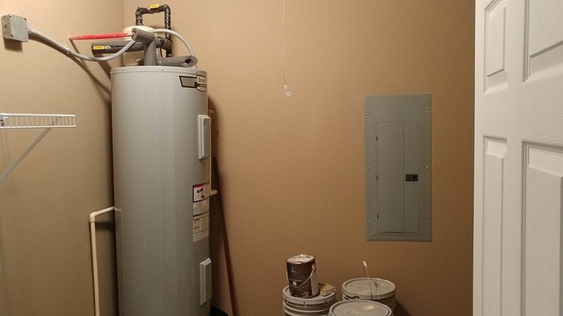 White cylindrical water heater with red labels positioned beside several plastic buckets, set against a bathroom wall with visible plumbing fixtures.