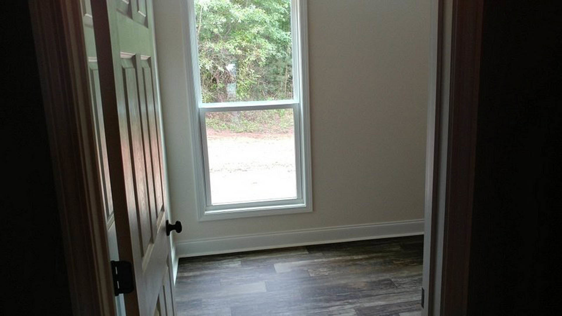 Sunlit room with large window overlooking trees, light wood flooring, white walls, and modern brushed metal door handle
