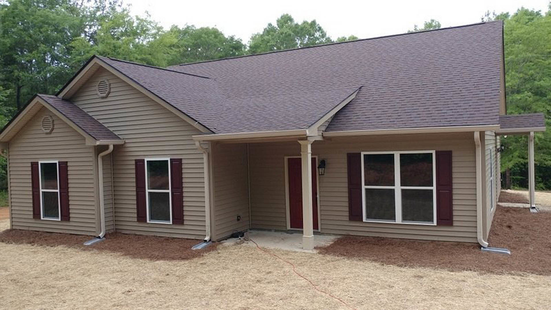 Brown shingle roof above white-framed windows, red and white siding, covered porch with entry door, wall vent, surrounded by trees