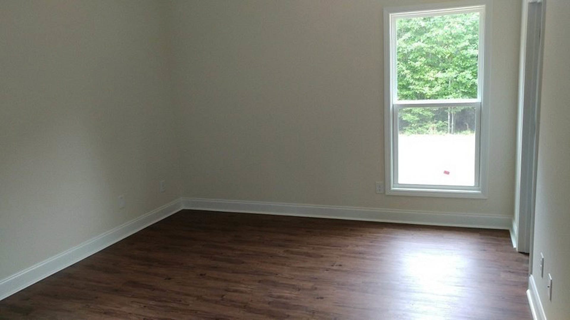 Wood flooring and white walls in a bright room with a large window, white curtain, and leafy trees visible outside