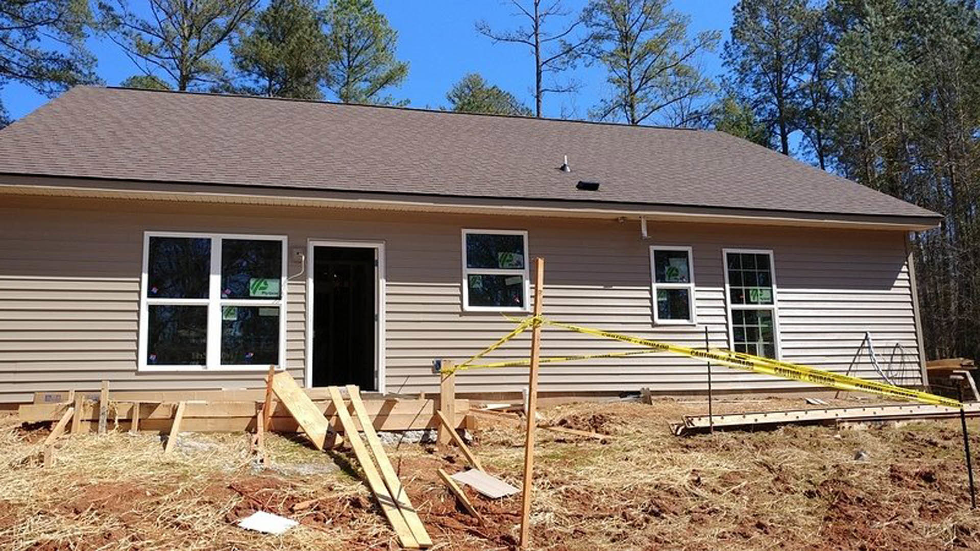 House under construction with yellow caution tape, white-framed windows, close-up of door, green and white logo on window, wooden planks scattered on ground, trees and sky in