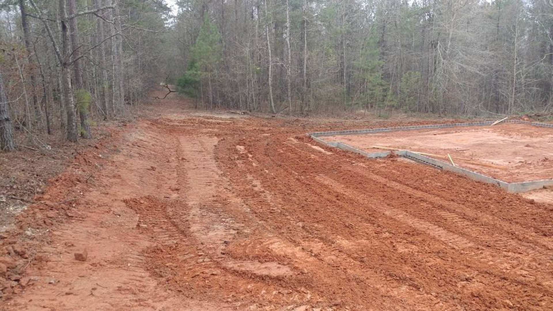 Dirt road bordered by trees, open soil field with square foundation, construction site materials, forested background