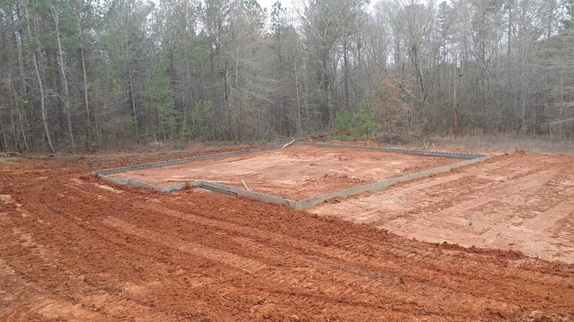 Concrete foundation slab surrounded by dirt and tire tracks, leafless trees in background, partial blurry person in foreground