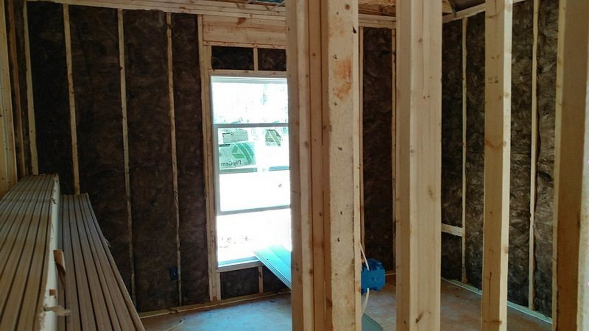 Bright room with large wood-framed window, natural hardwood walls, and visible plywood beams; blue plastic container with white straw sits near window.
