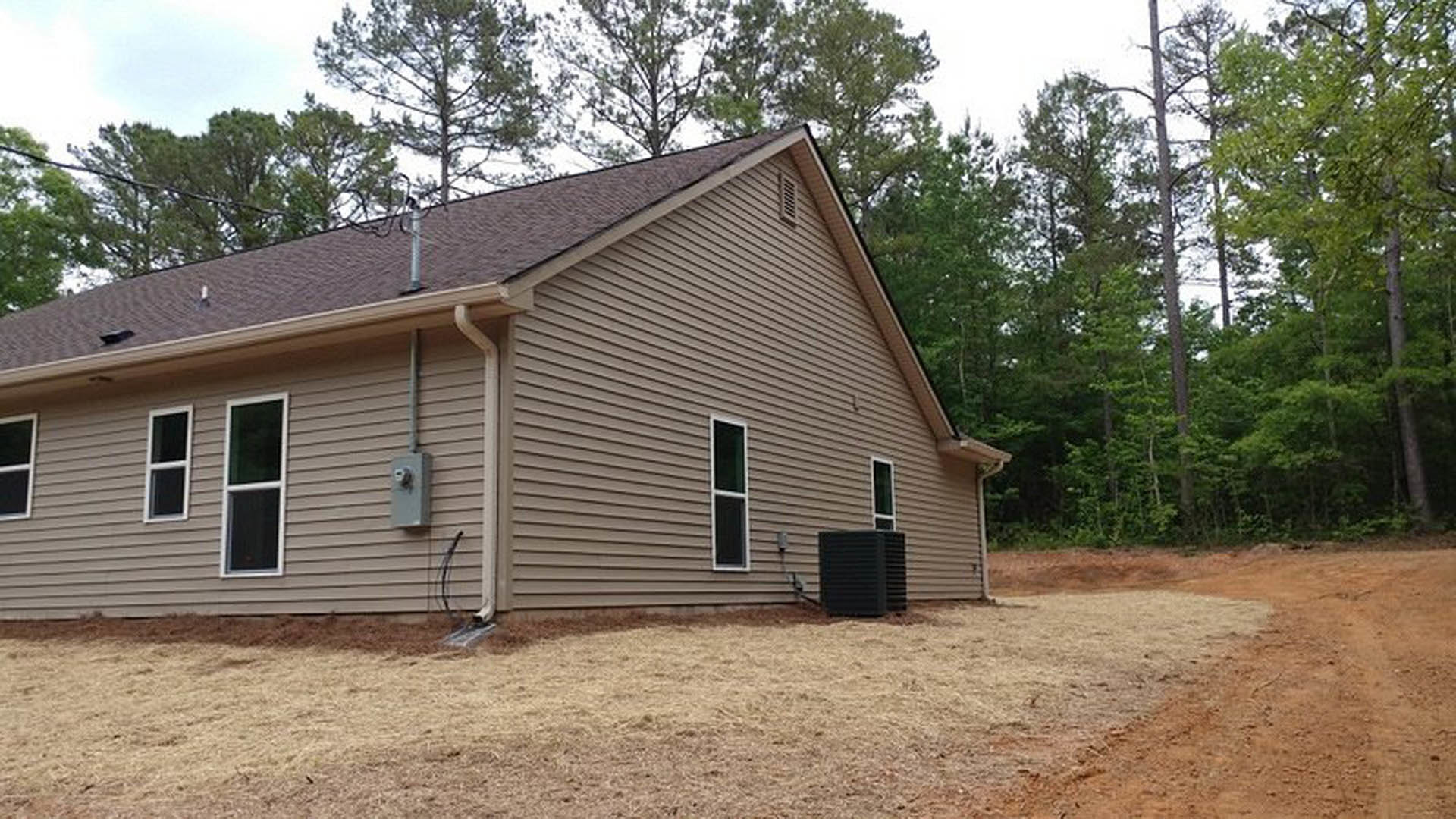 Two-story home with light siding, white-framed windows, black utility box, visible gutter, dirt driveway, and surrounding mature trees