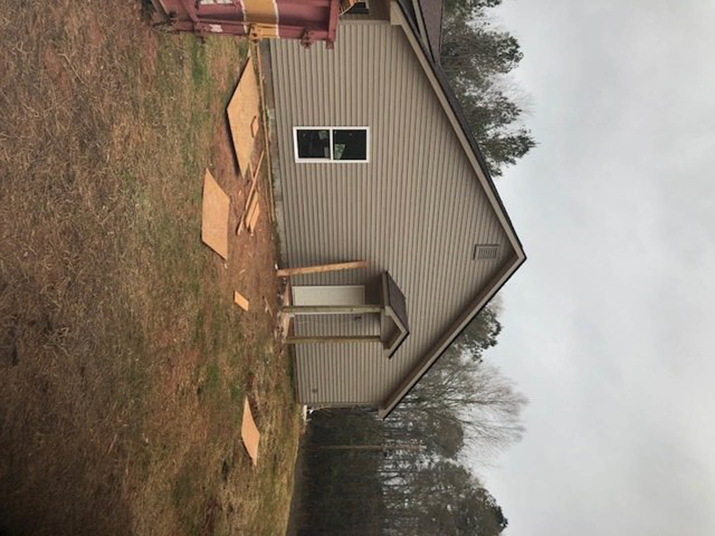 Two-story cottage with white-framed windows, fenced grassy yard, scattered wood pieces, metal beam, and mature trees under cloudy sky