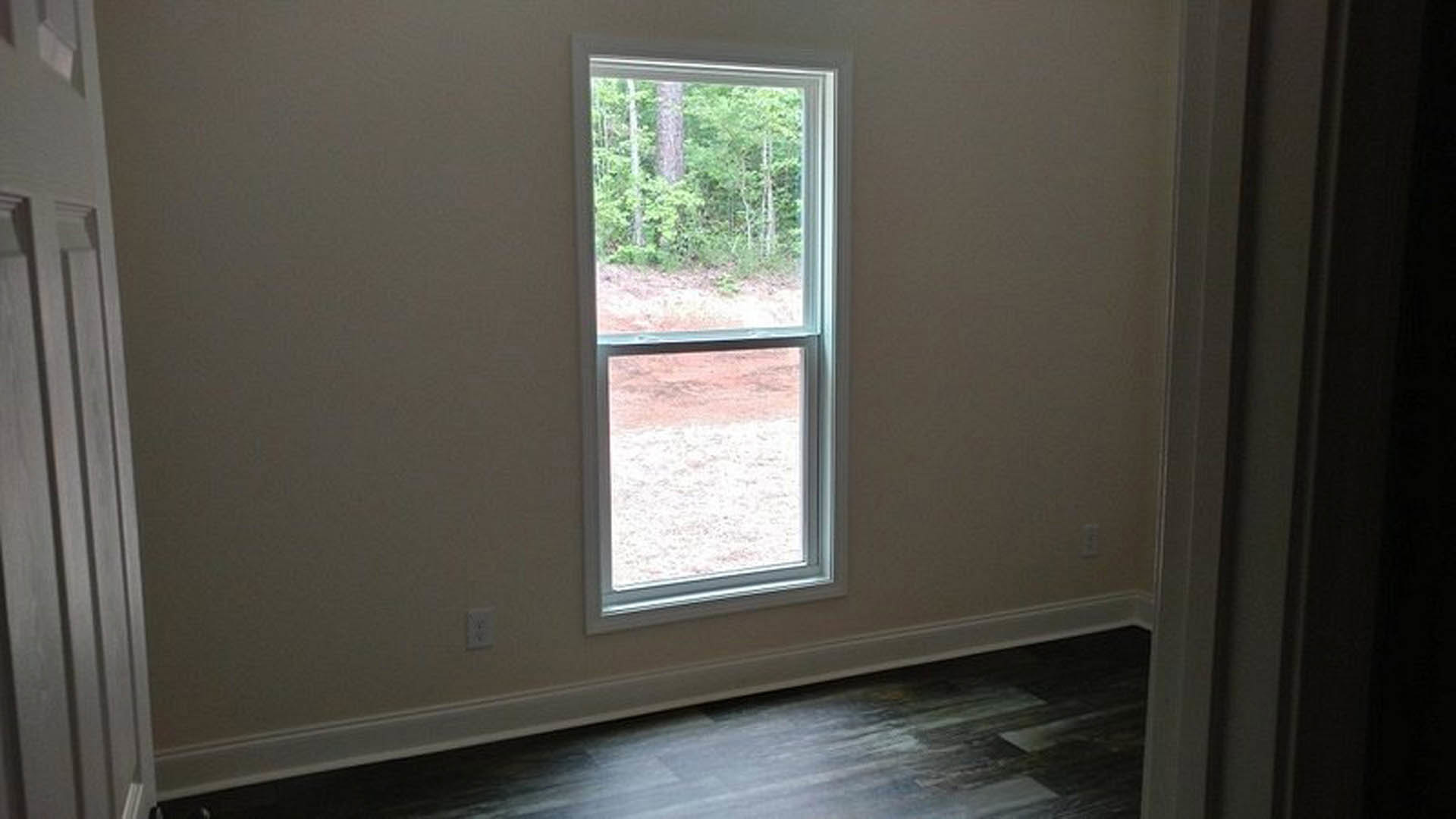 Large window with white trim overlooking leafy trees, dark wood floor with white baseboard, white electrical outlet on pale wall, partial view of white door