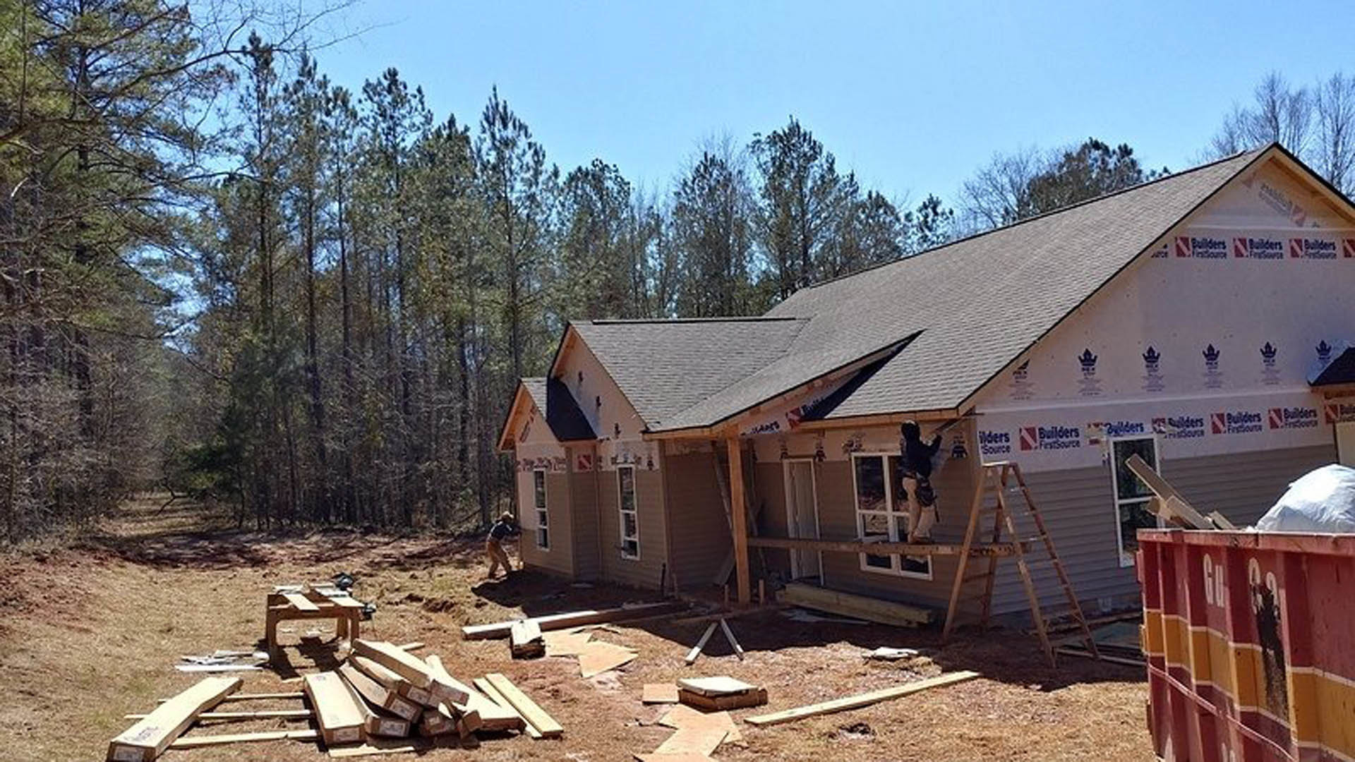 Wood-framed house under construction surrounded by tall trees, exposed lumber walls, man working on ladder, red and yellow dumpster nearby on dirt ground