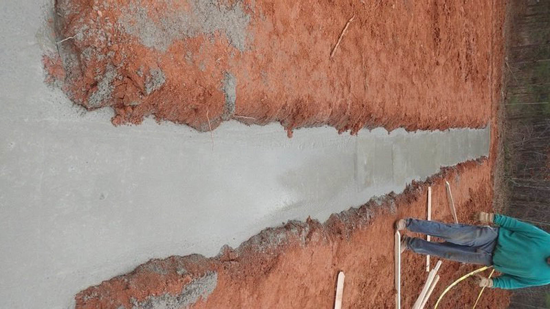 Man in green shirt and blue jeans holding yellow hose standing on dirt next to concrete slab outside custom home with painted exterior walls