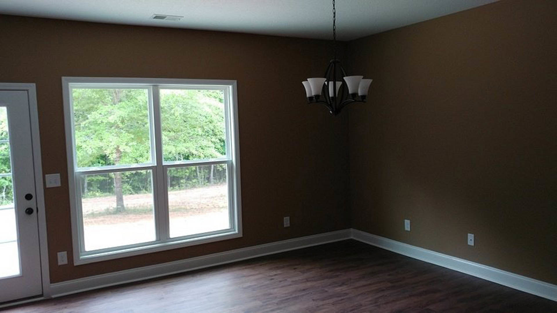 Crystal chandelier hanging from ceiling above hardwood floor, large window with leafy trees outside, white walls, modern door handle in foreground
