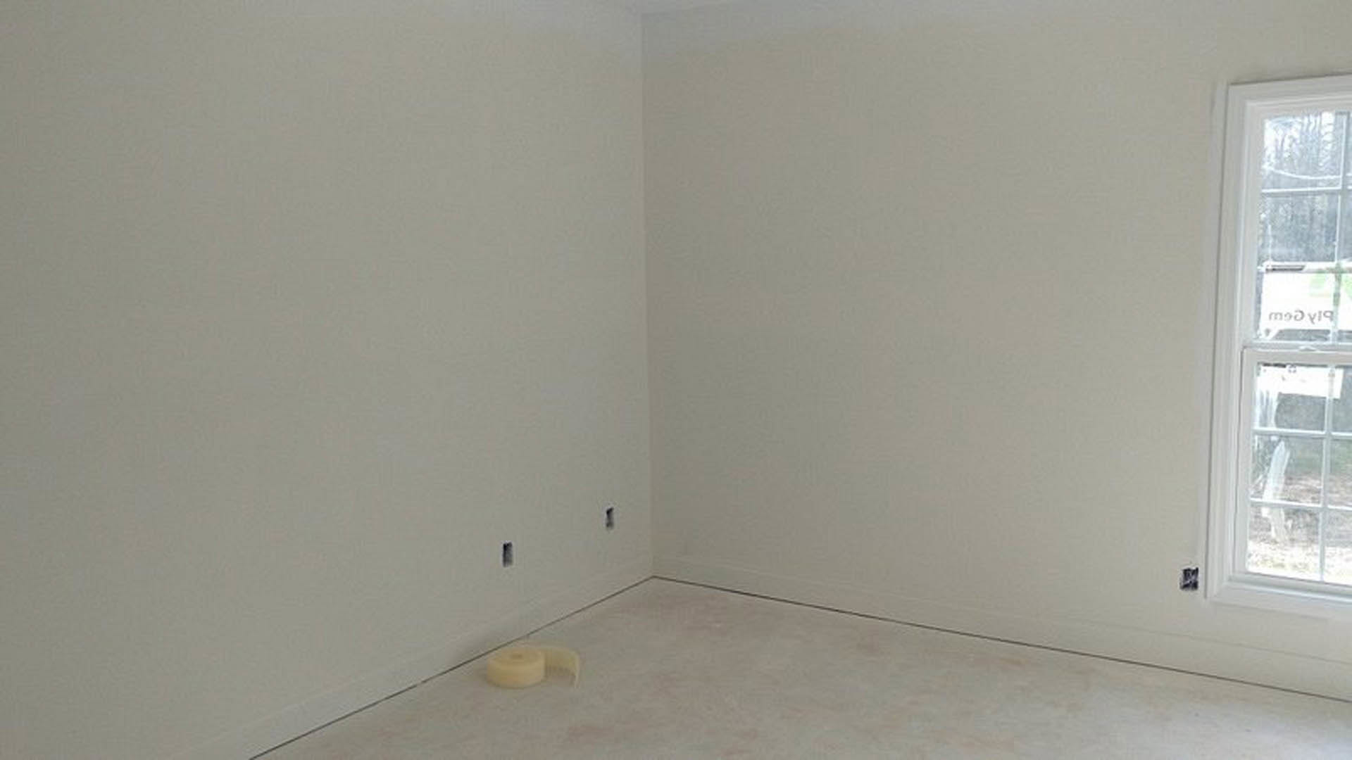 Unfinished room with concrete floor, white plaster walls, laminate flooring in progress, roll of tape resting on a table near window with sign
