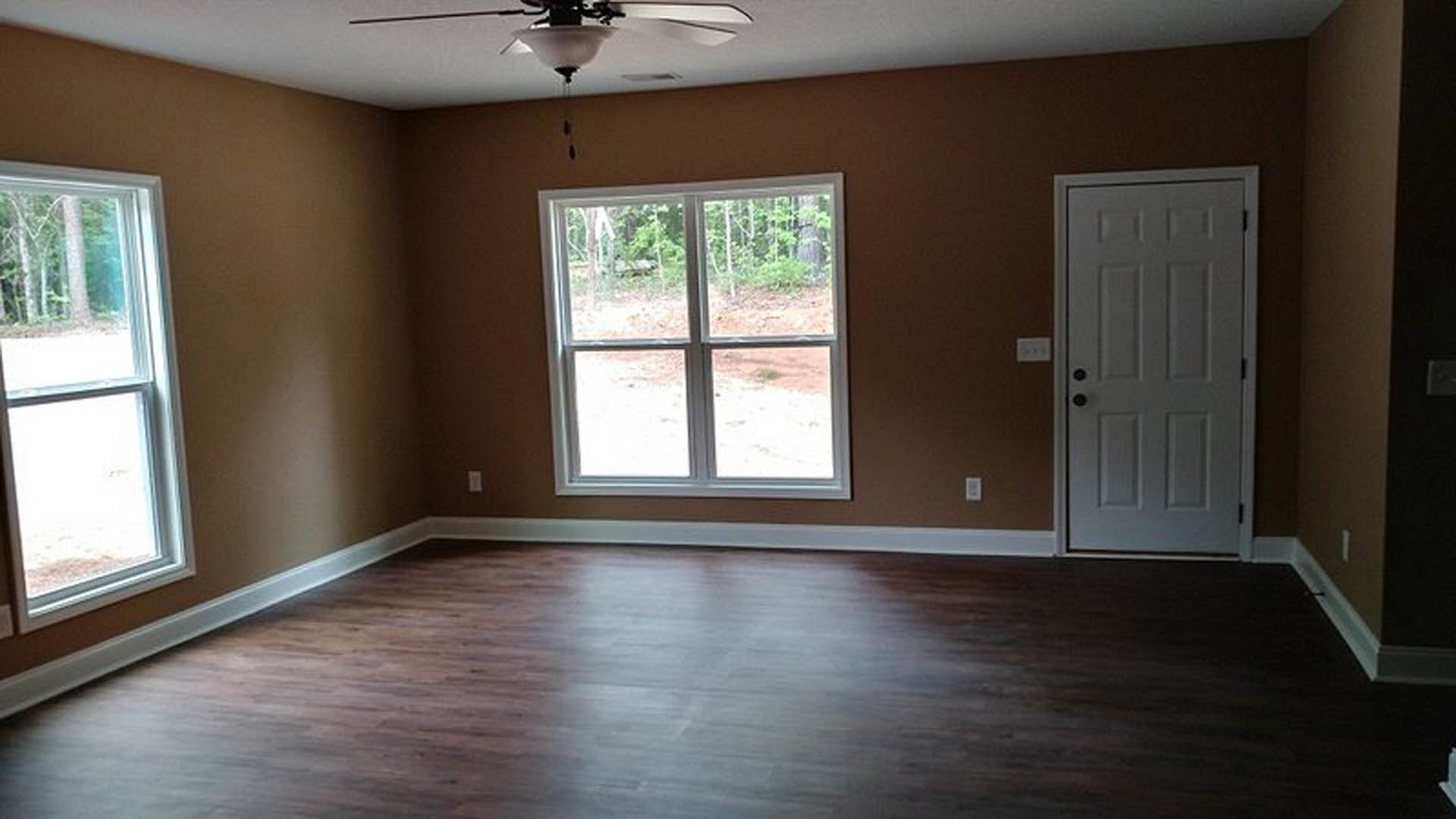 Wood flooring with white baseboards, white door featuring black knobs, window overlooking forested trees, ceiling fan with light fixture
