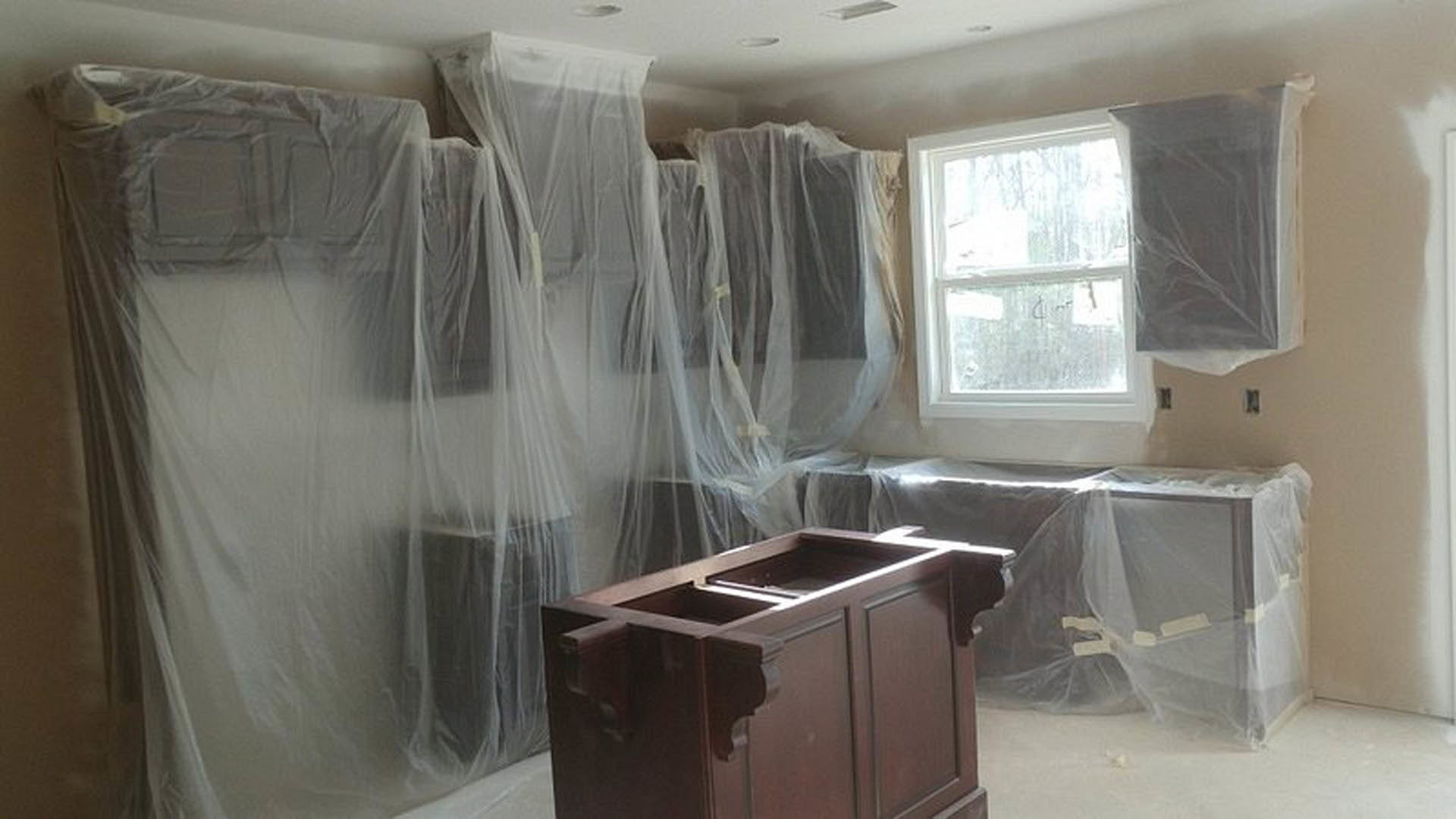 Room under renovation with plastic coverings draped over a table and window, wooden cabinet with drawers, white-framed window, white rectangular object on a white surface.