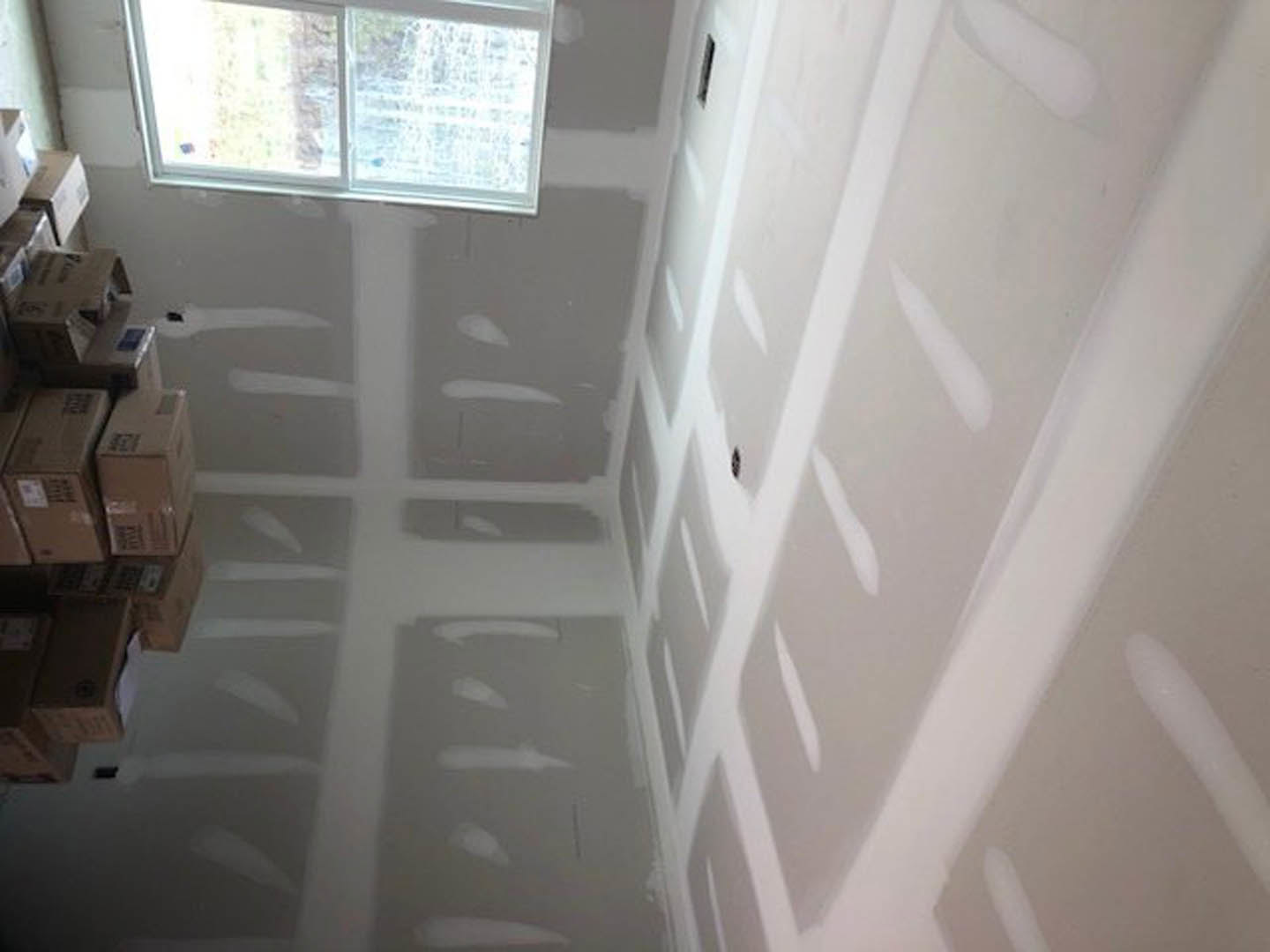 Bright room with smooth white plaster walls, large window letting in daylight, light wood floor, and stacked moving boxes near the corner