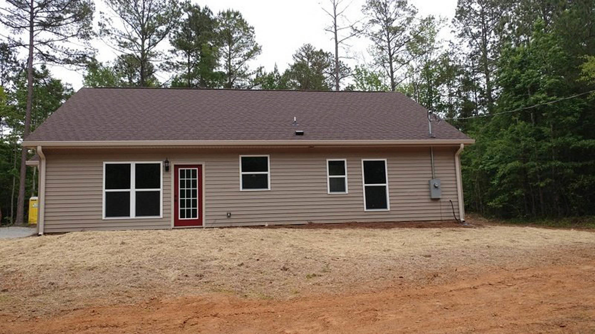 Two-story cottage with gray siding, square white-trimmed windows, and a prominent red front door, set on a grassy dirt yard with trees in the background.