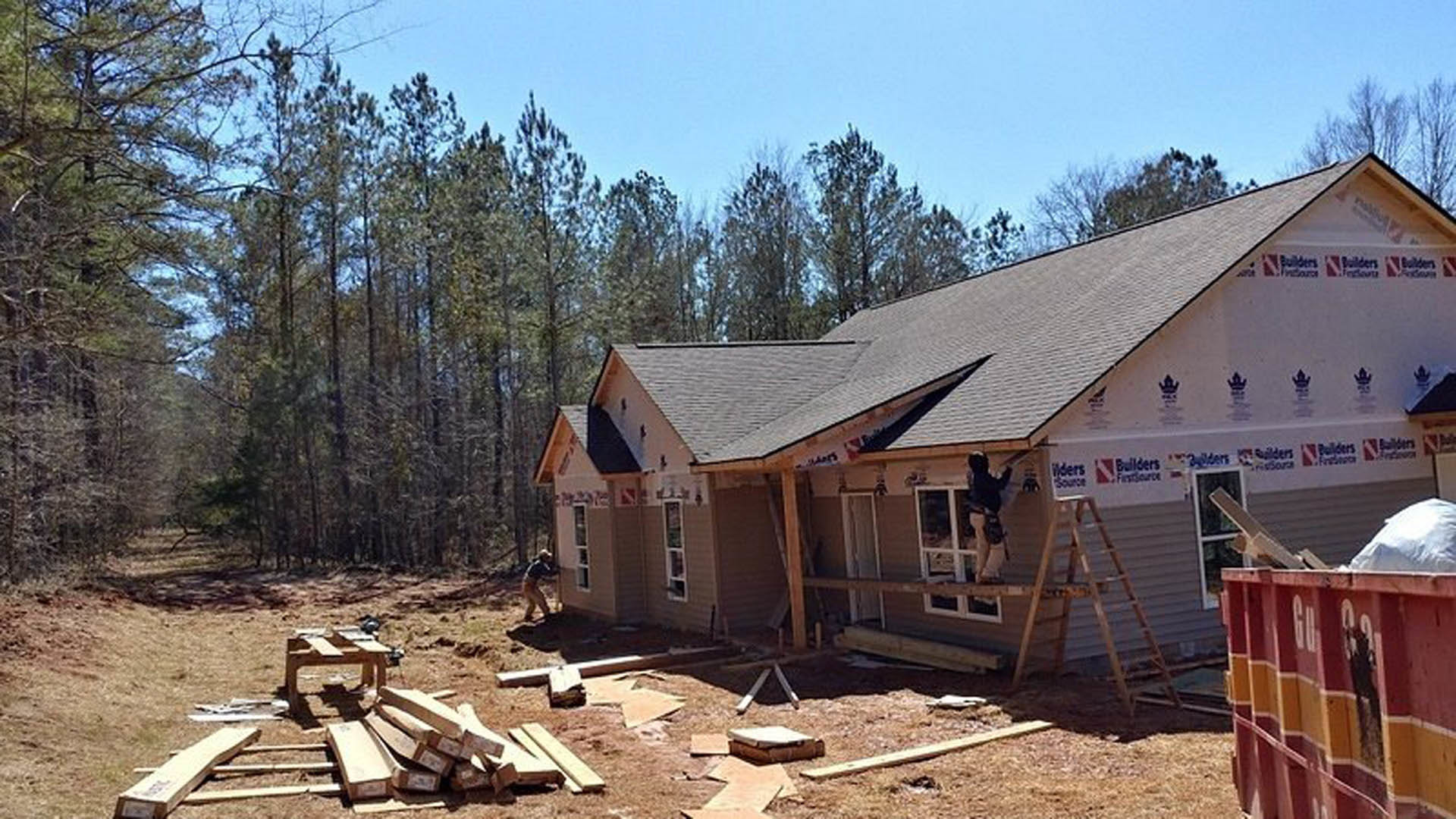 Wood-framed house under construction surrounded by tall trees, person on ladder working near exterior, red and yellow dumpster and cardboard boxes scattered on dirt ground