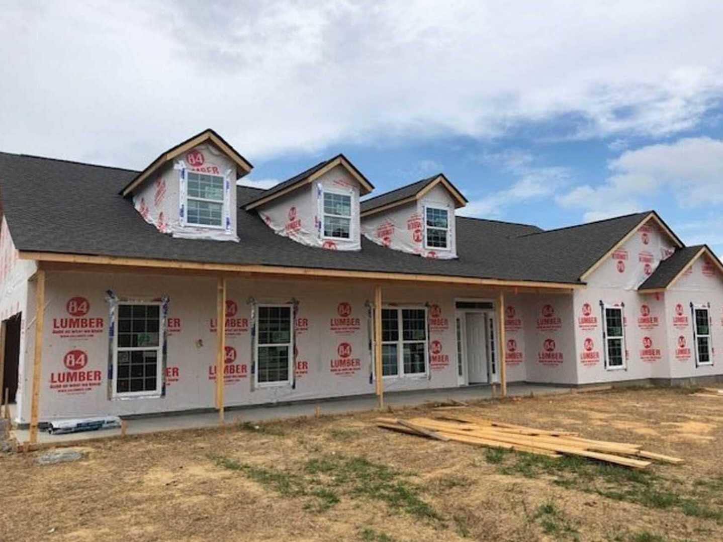 Partially built house with white-framed windows, red and white construction signs, exposed wood framing, piles of lumber on dirt, gray siding, and a shingled roof under a blue sky.