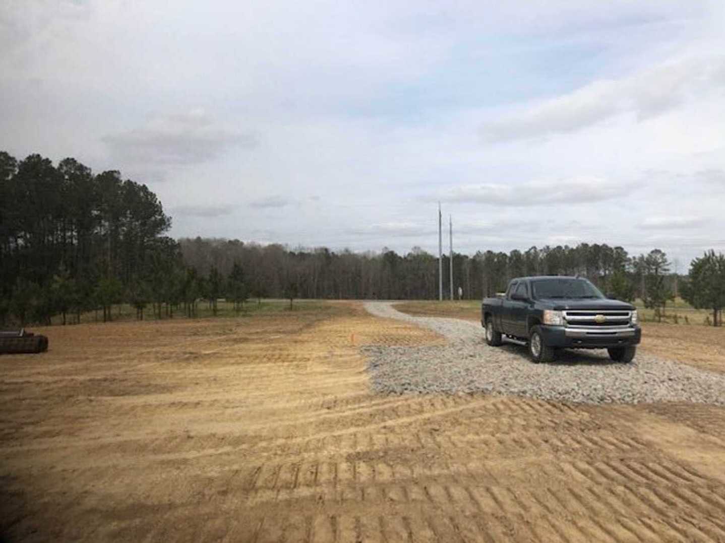 Black pickup truck parked on a gravel driveway beside a dirt area, tire tracks visible in the foreground, cloudy sky overhead, trees lining the background.