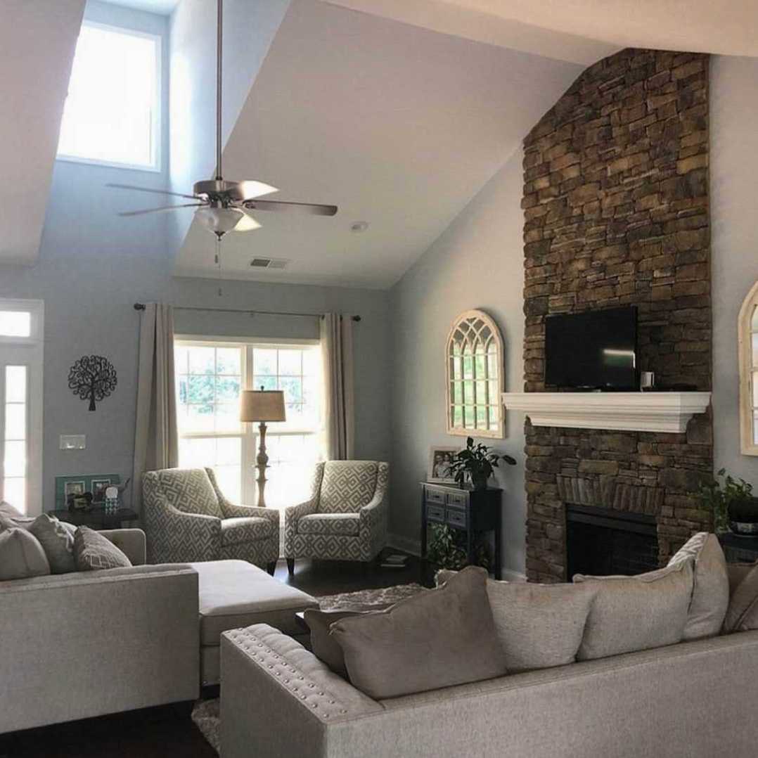 Living room with brick fireplace, gray couches with patterned pillows, side table with lamp near large multi-pane windows, decorative black metal wall art with swirling tree