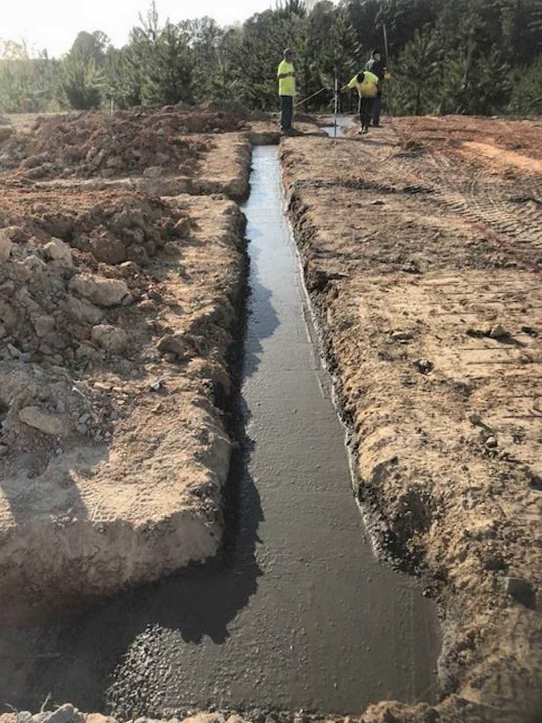Man standing beside a dirt ditch with scattered rocks and a pipe, surrounded by trees and soil under an open sky