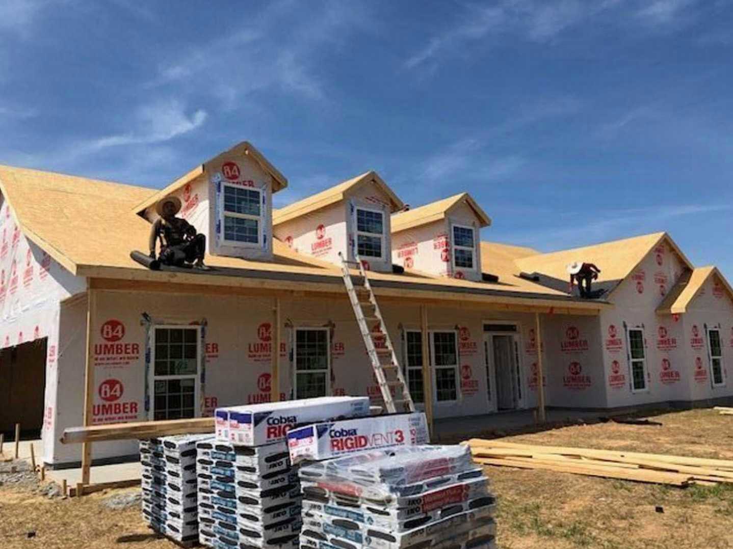 Shingle roof with a man in a hat standing near a ladder, cloudy sky above, house exterior with windows visible