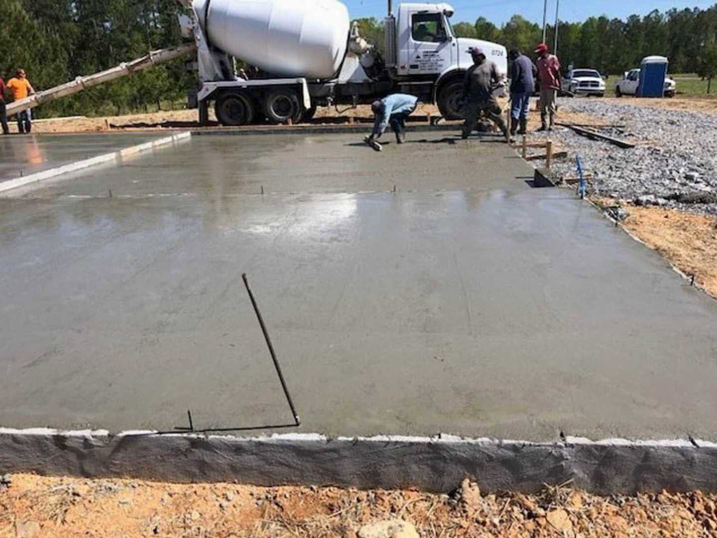 Men gathered beside freshly poured concrete slab, cement truck in background, outdoor setting with trees and sky visible