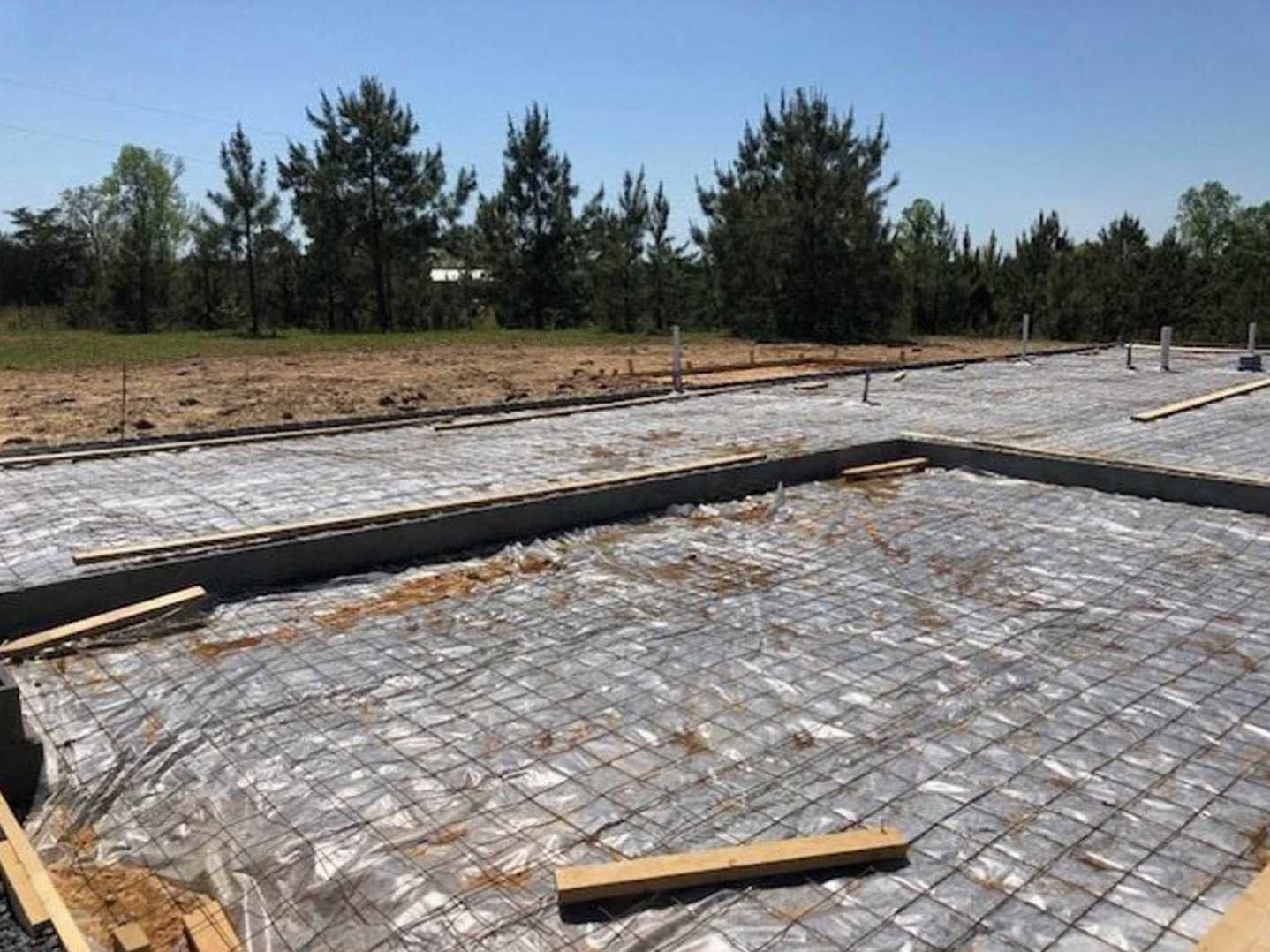 Framed custom home under construction with exposed metal grid and wooden planks, surrounded by trees and a dirt road with fencing in the background