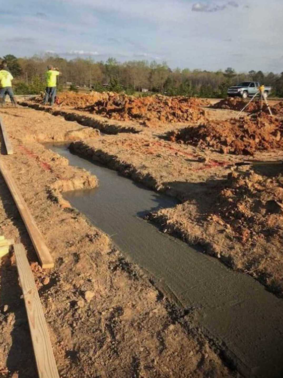 Framed custom home under construction with workers on site, wooden planks scattered on soil, trees and cloudy sky in background, construction truck partially visible