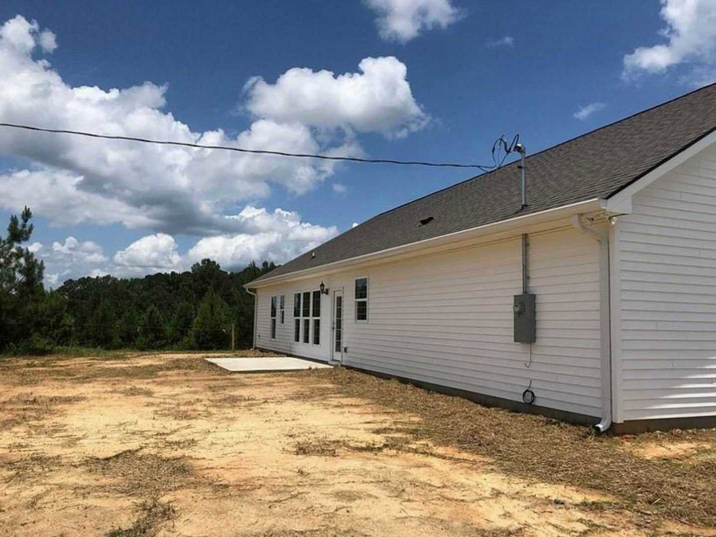 White house with black roof, multiple windows, dirt yard in foreground, scattered trees and cloudy sky in background