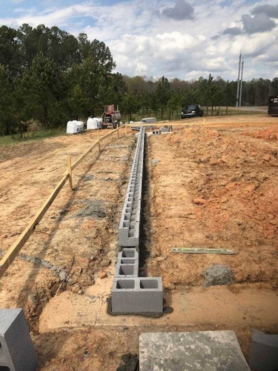 Concrete block wall under construction on a grassy site with soil, scattered plants, and cloudy sky in the background