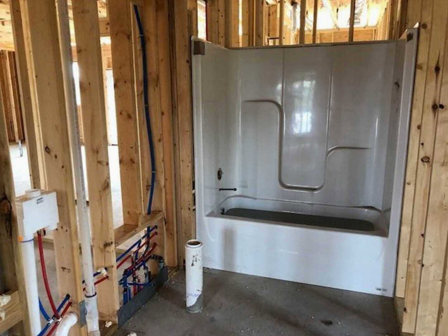 Bathroom with freestanding white tub, walk-in shower, exposed concrete floor, unfinished plaster walls, visible lumber beams, and building insulation.