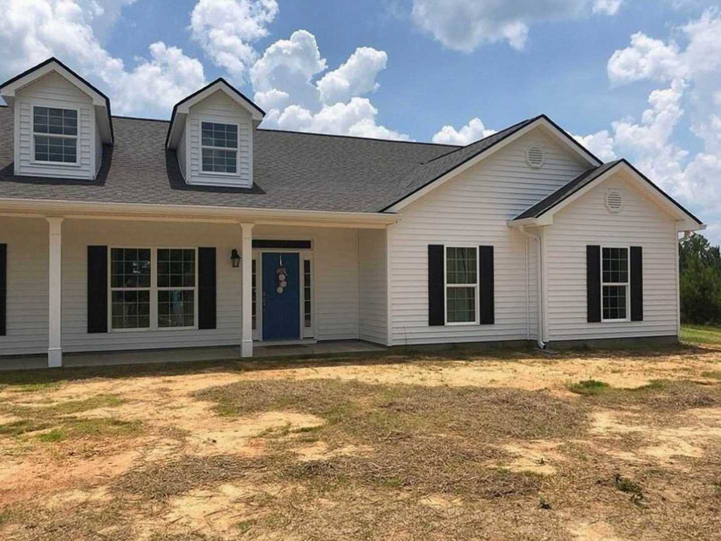 Blue front door with white decorative knobs, multi-pane window with white frame, gray siding, patch of green grass, partly cloudy blue sky.