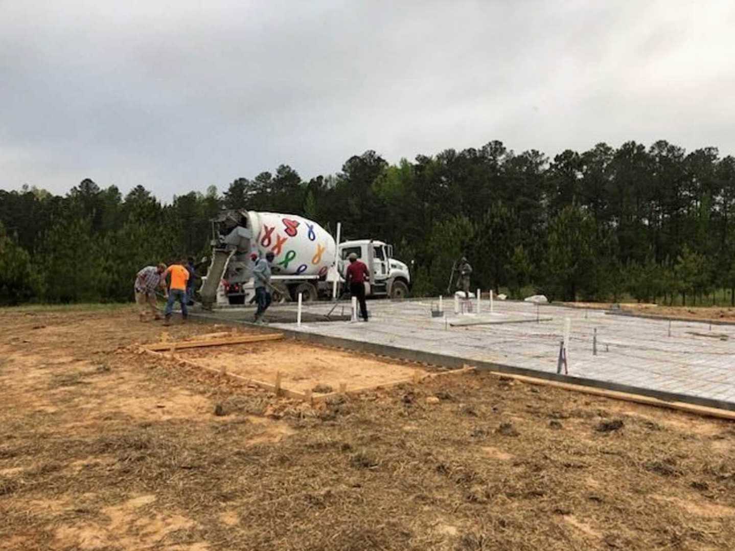 Group of people standing on dirt ground beside a concrete mixer and white truck, with trees and sky in background