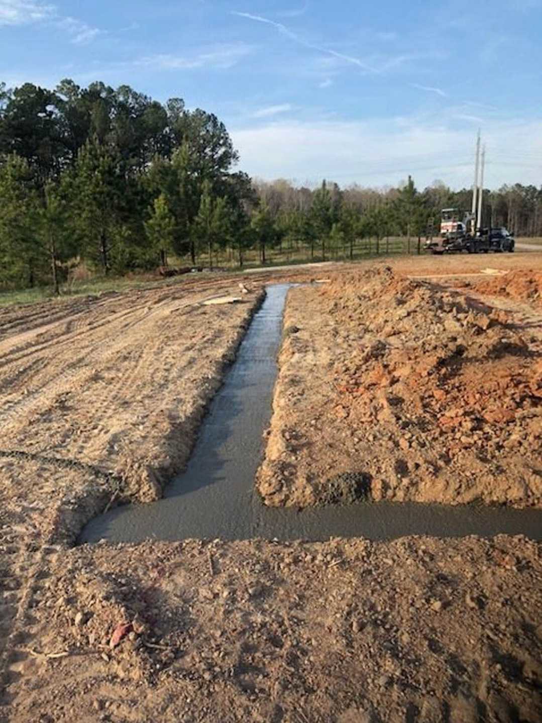 Drainage ditch with water running through a dirt field, bordered by trees under a blue sky, tractor parked nearby