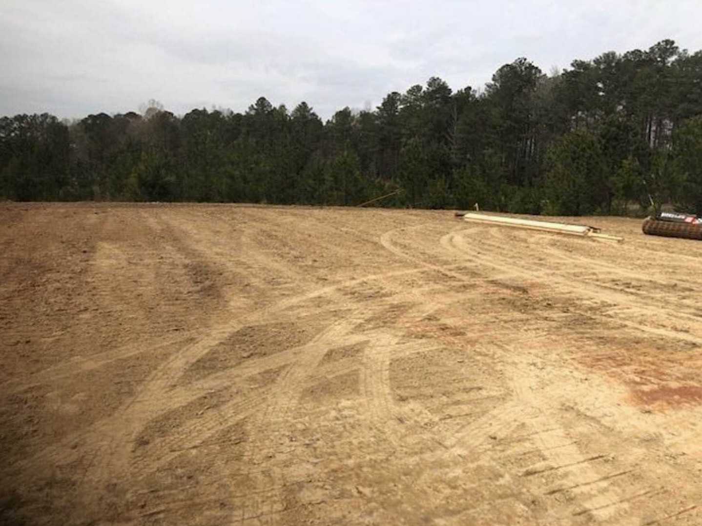 Dirt field with tire tracks, scattered trees in the background, cloudy sky overhead, and a blurred vehicle near the edge of the lot