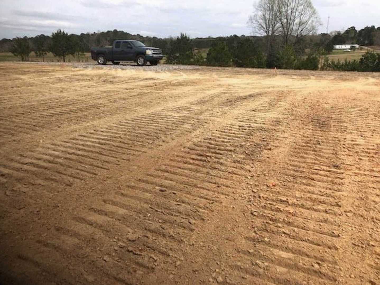 Black pickup truck parked on a dirt driveway beside a grassy field, surrounded by dense trees under a cloudy sky