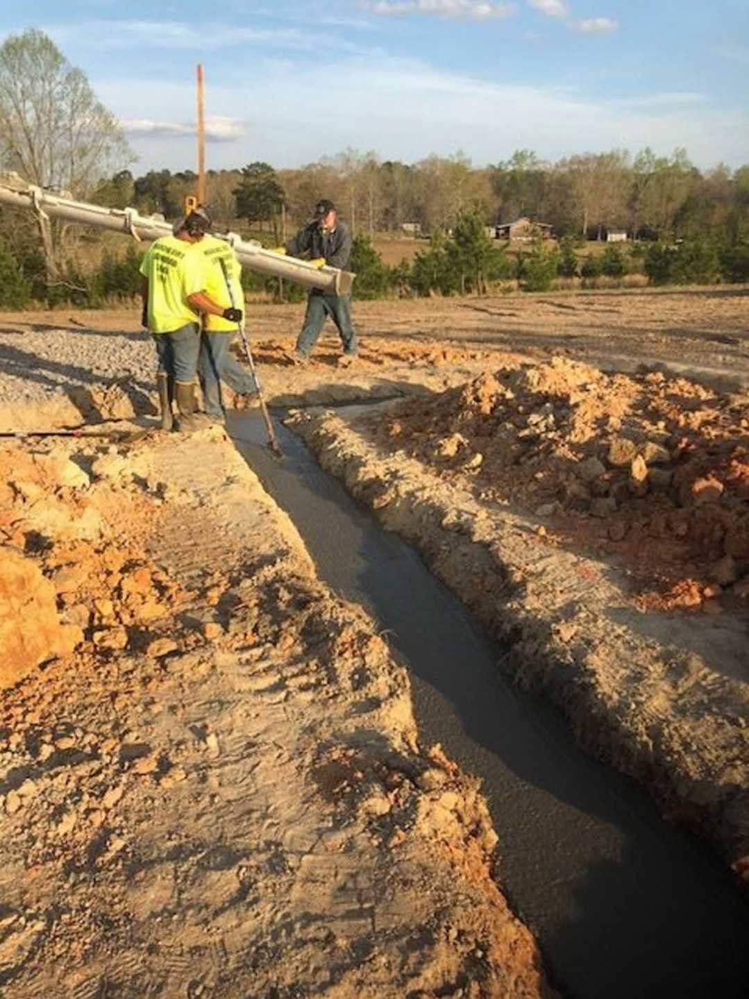 Four men working outdoors on a residential construction site, one carrying a metal pipe, others wearing yellow shirts and gloves, with exposed soil, a ditch, and trees in the
