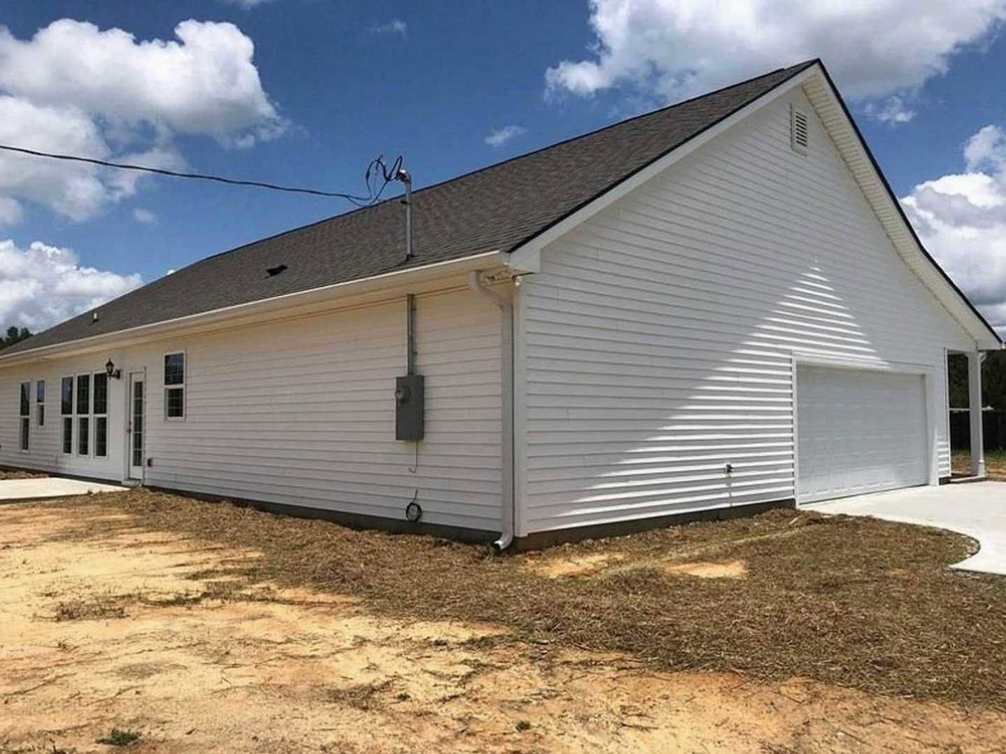 White siding home with attached garage, multiple windows, gray shingle roof, and dirt landscaping under a partly cloudy sky.