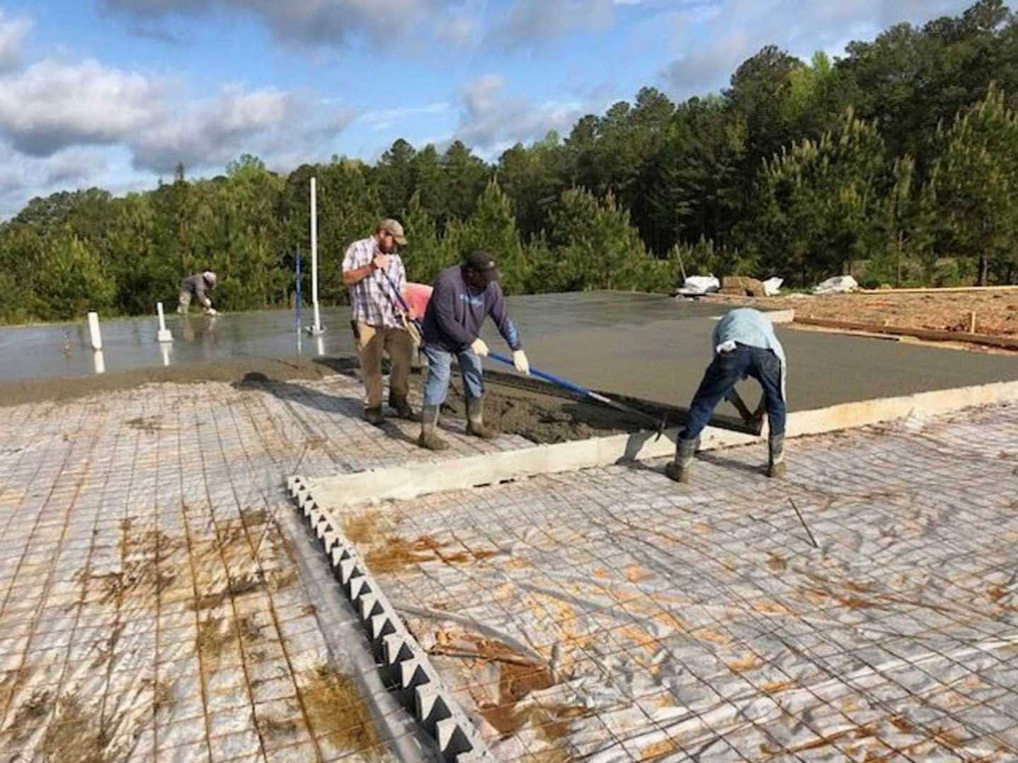 Workers pouring and leveling fresh concrete slab outdoors, surrounded by construction materials, trees, and cloudy sky