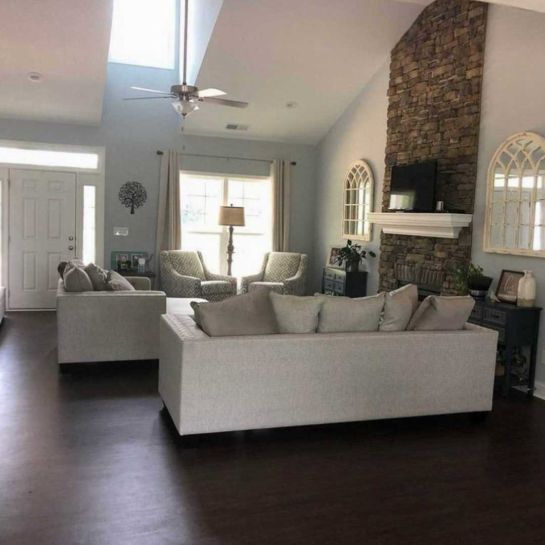 Living room with gray couch and patterned pillows, stone fireplace, metal tree wall art, large window, wood flooring, ceiling fan with light fixture