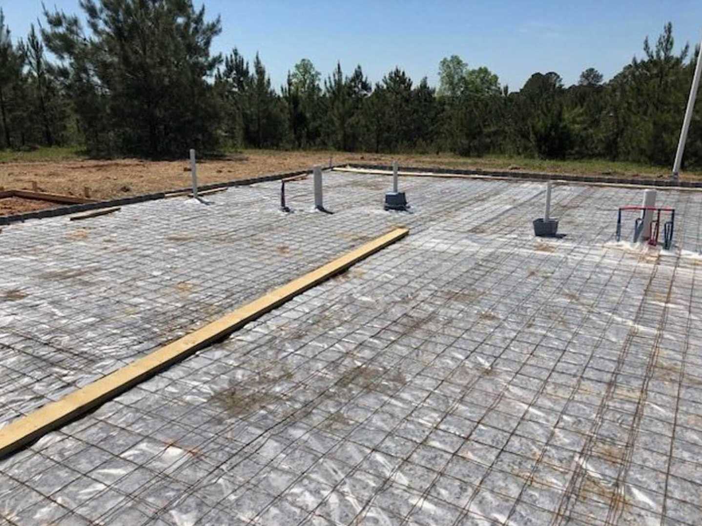 Concrete slab foundation with exposed metal rebar, surrounded by trees and construction materials on a dirt lot