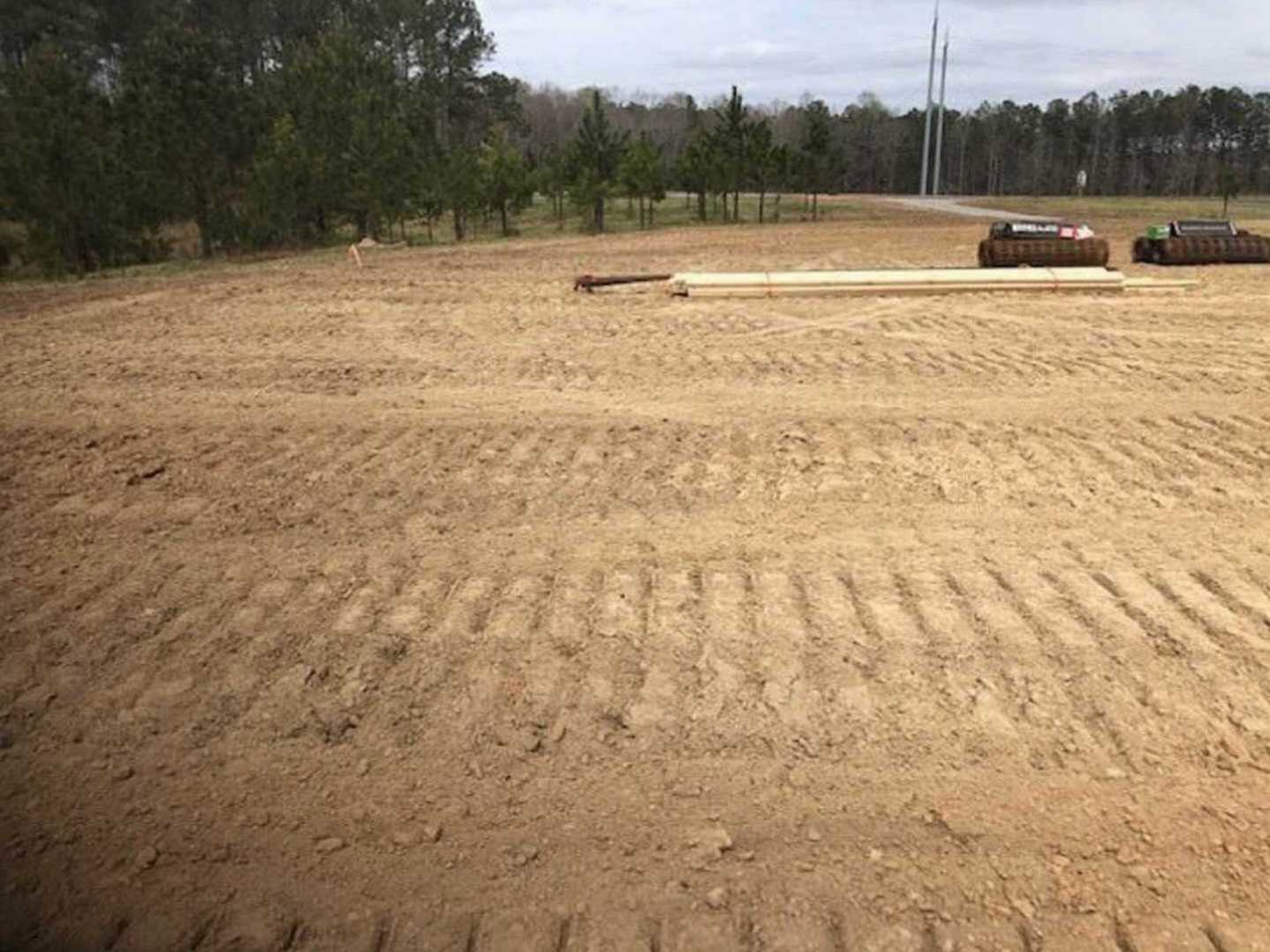 Dirt field with tire tracks, tractor parked near tall poles, sparse trees in background under open sky